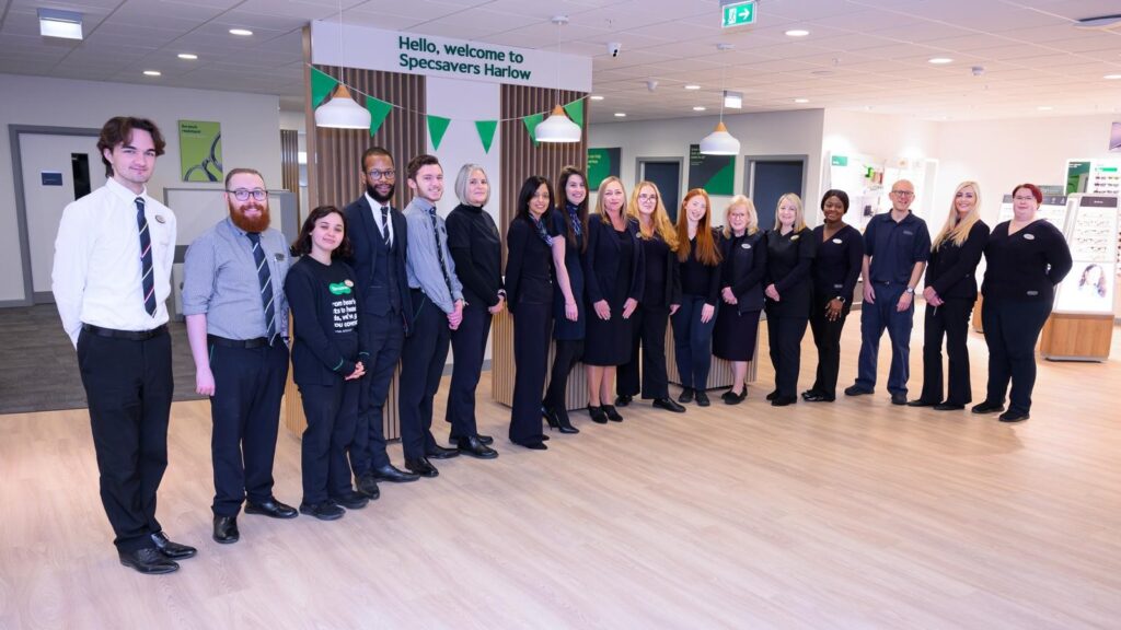 A group of staff standing together under a sign which says Hello, welcome to Specsavers Harlow and has green bunting flags underneath it