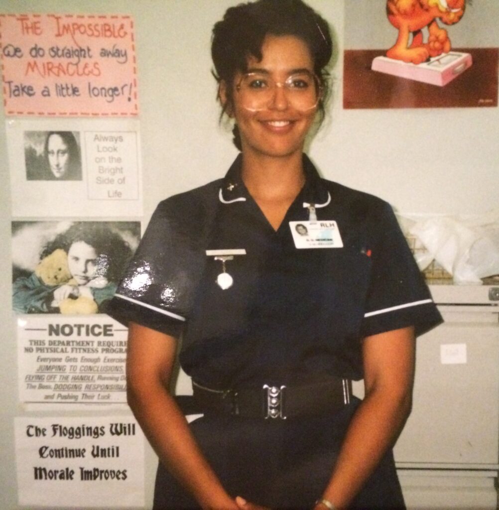 Sharon Hodgson standing in front of a wall with pictures on it, wearing a Navy Blue nurse's uniform.