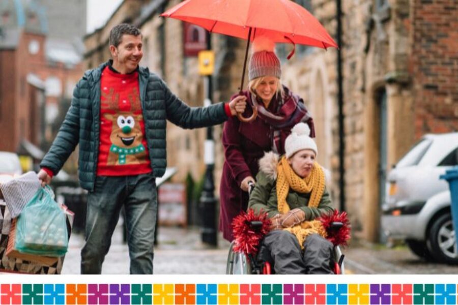 A man and a woman and a child in Christmas jumpers and wrapped up warm laughing in the street with snow falling along the street. Man holding an umbrella. Young girl is in a wheelchair