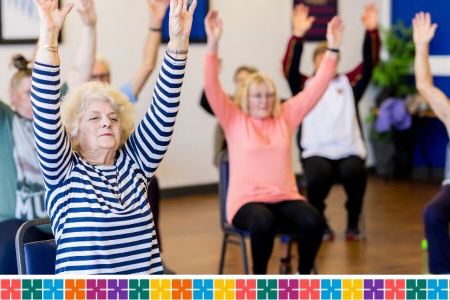 Group of older people sitting on chairs in a hall with hands in the air. They are taking part in a seated exercise.