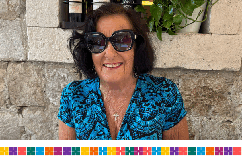 An elderly woman with dark hair and wearing sunglasses and a blue dress in front a a limestone wall.