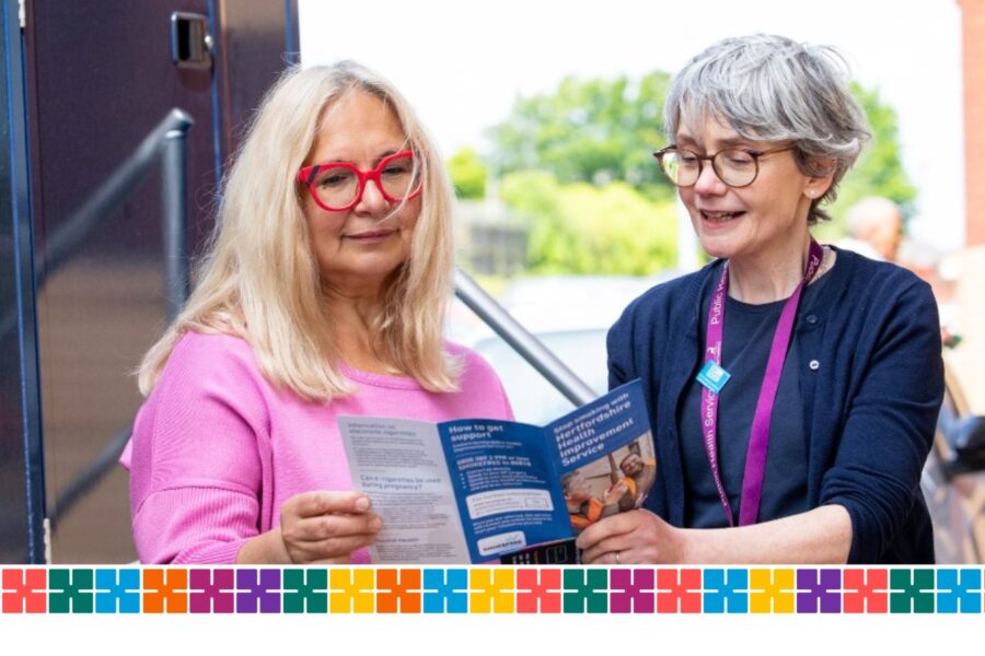 Two women looking at a bus time table together, one wearing a pink top, he other a blue one and both with glasses