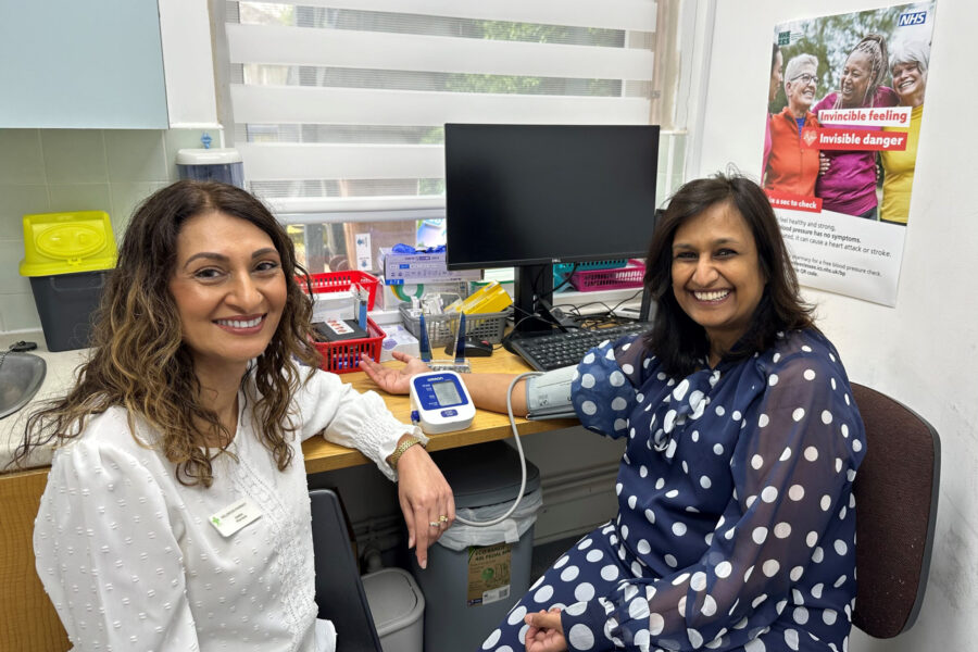 Two women smiling sitting in an office with one having a blood pressure cuff on her arm and the blood pressure machine on the desk behind along with a computer screen